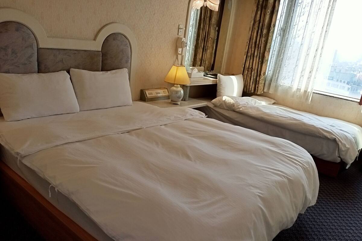 Une chambre d’hôtel avec un grand lit, un lit plus petit, une table de chevet avec une lampe et une fenêtre avec des rideaux.
