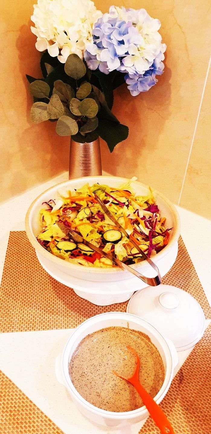 A bowl of salad with a spoon, a small bowl of soup with a spoon, and a vase with flowers.