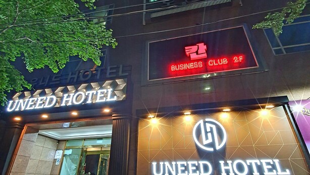 Ein Hotel mit einem gut sichtbaren Schild 'UNEED HOTEL', einem Schild 'BUSINESS CLUB 2F' und einem Schild 'HOTEL' über dem Eingang.