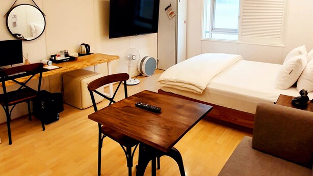 A modern, well-lit room with a wooden floor, a bed, a sofa, a desk with a chair, a television, and a ceiling light fixture.