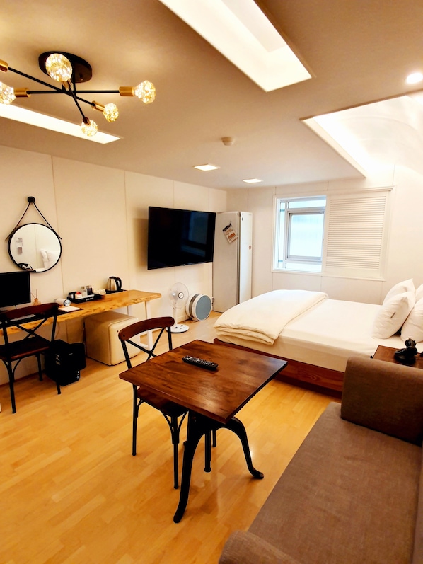 A modern, well-lit room with a wooden floor, a bed, a sofa, a desk with a chair, a television, and a ceiling light fixture.