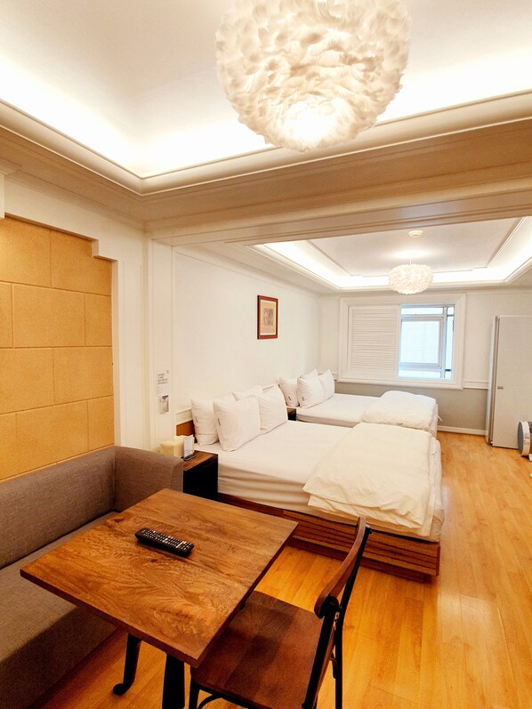 A modern hotel room with a wooden floor, a sofa, a bed with white linens, a wooden table with a remote control, and a ceiling light fixture.