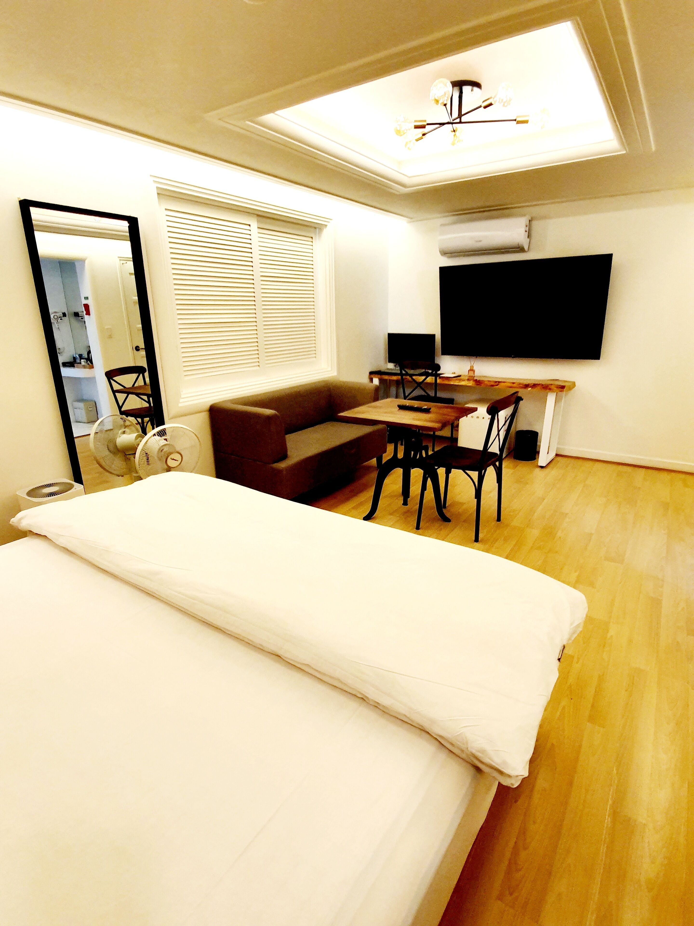 Quarto de hotel compacto com cama, sofá, escrivaninha com cadeira, TV e ventoinha de teto.