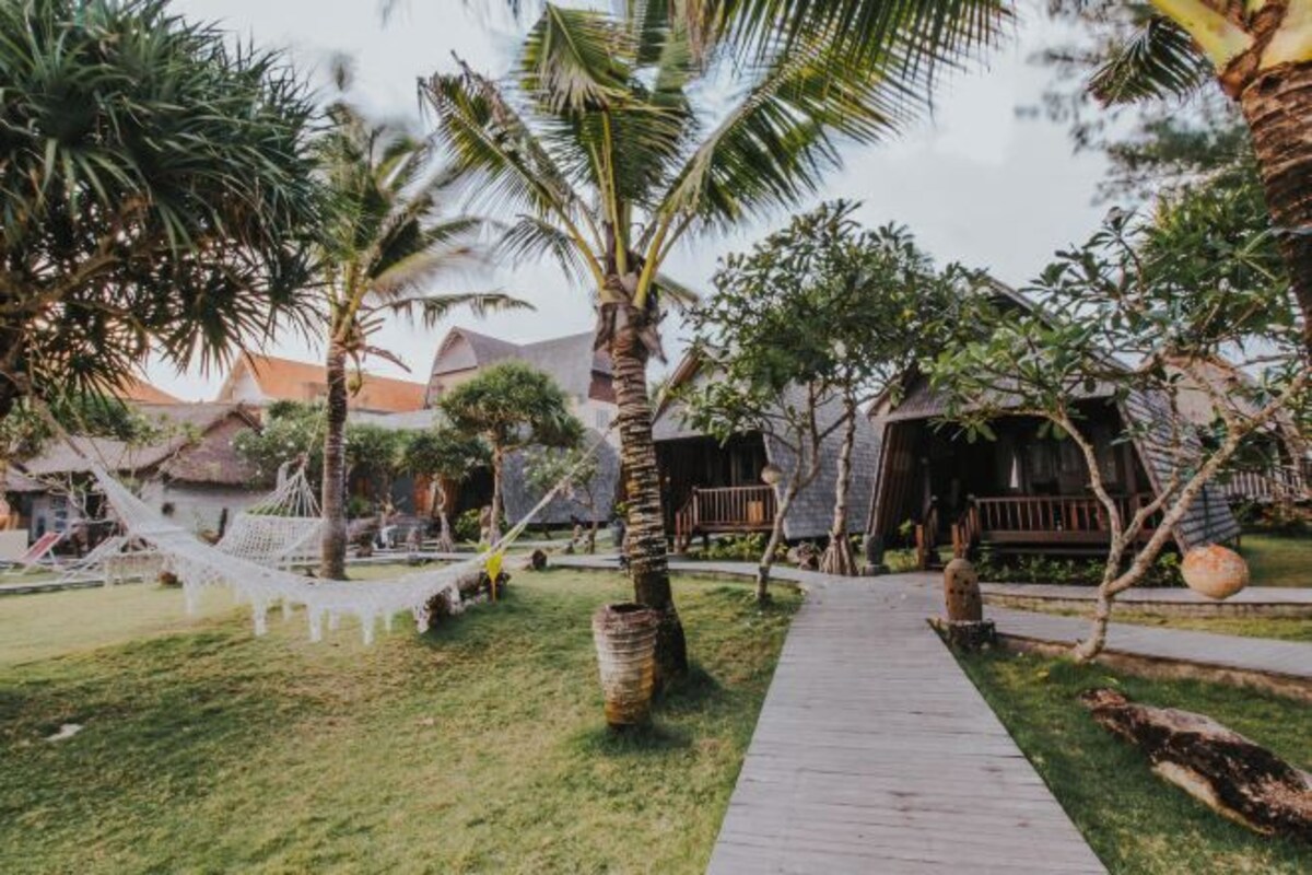 A wooden pathway leading to a building with a thatched roof, surrounded by palm trees and a hammock.