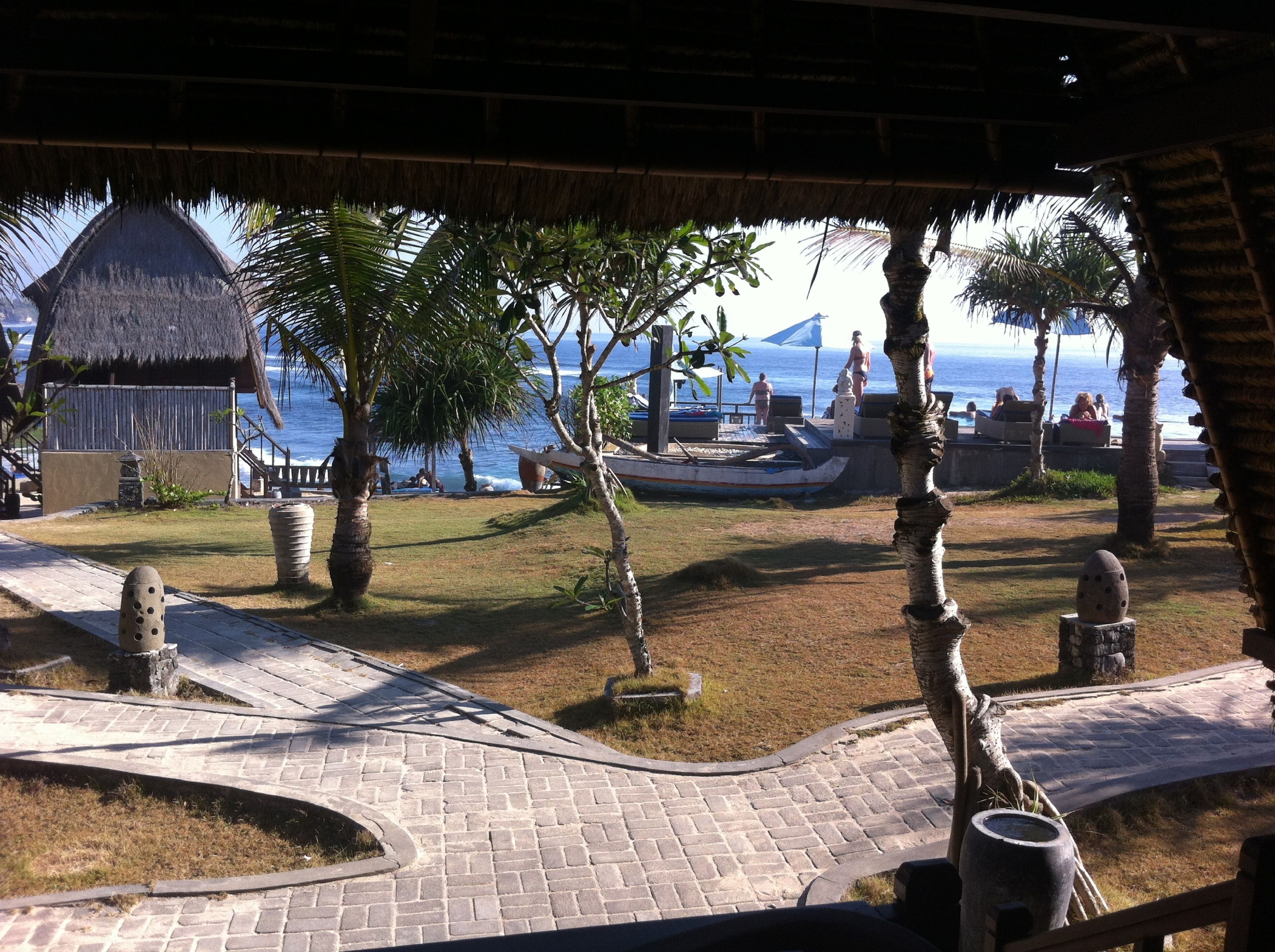 A tropical resort with a thatched-roof hut, palm trees, a boat, and a paved pathway leading to the beach.