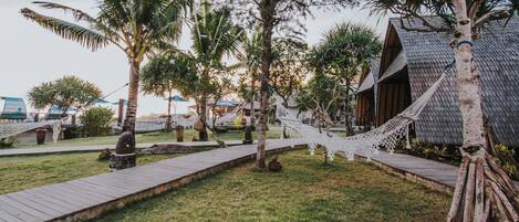 A wooden pathway leading to a building with a thatched roof, surrounded by palm trees and a hammock.
