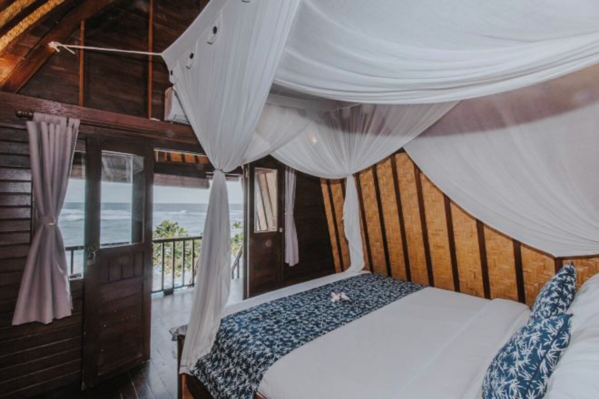 A bedroom with a bed, white curtains, and a view of the ocean.
