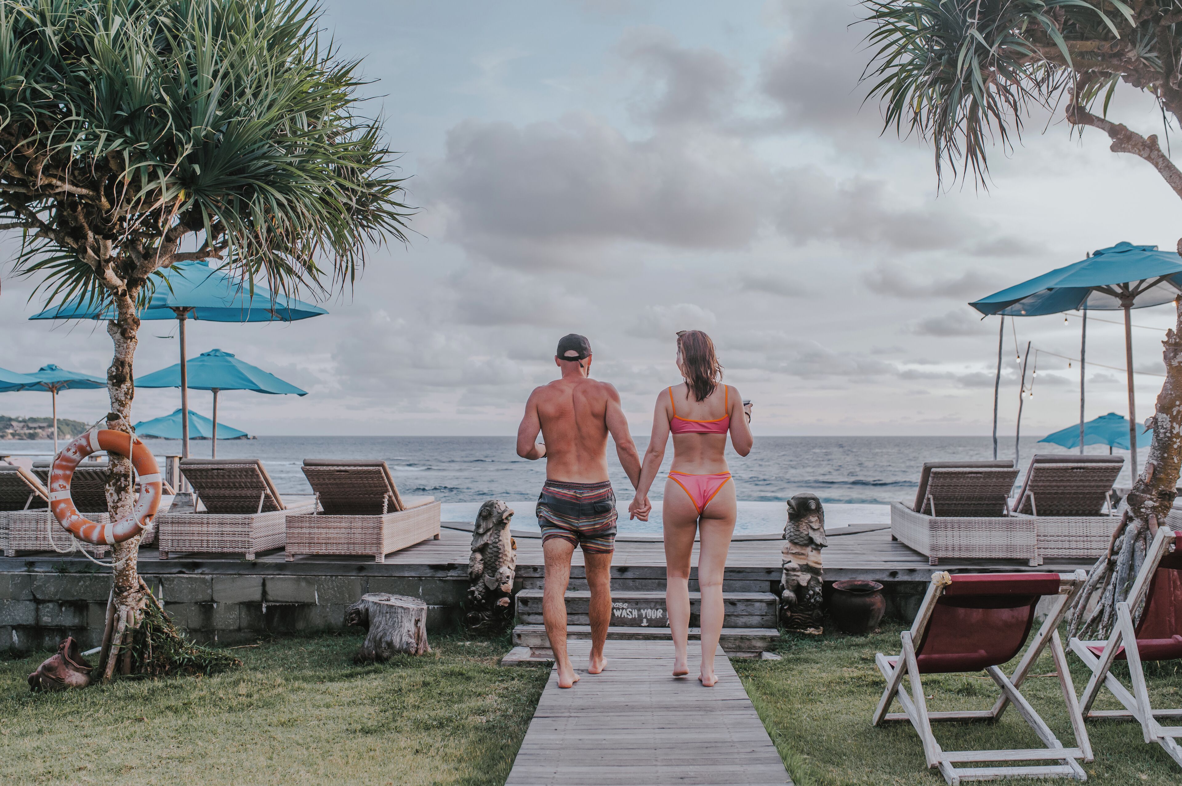 A couple walking on a wooden deck towards the ocean, with lounge chairs and umbrellas on the side.