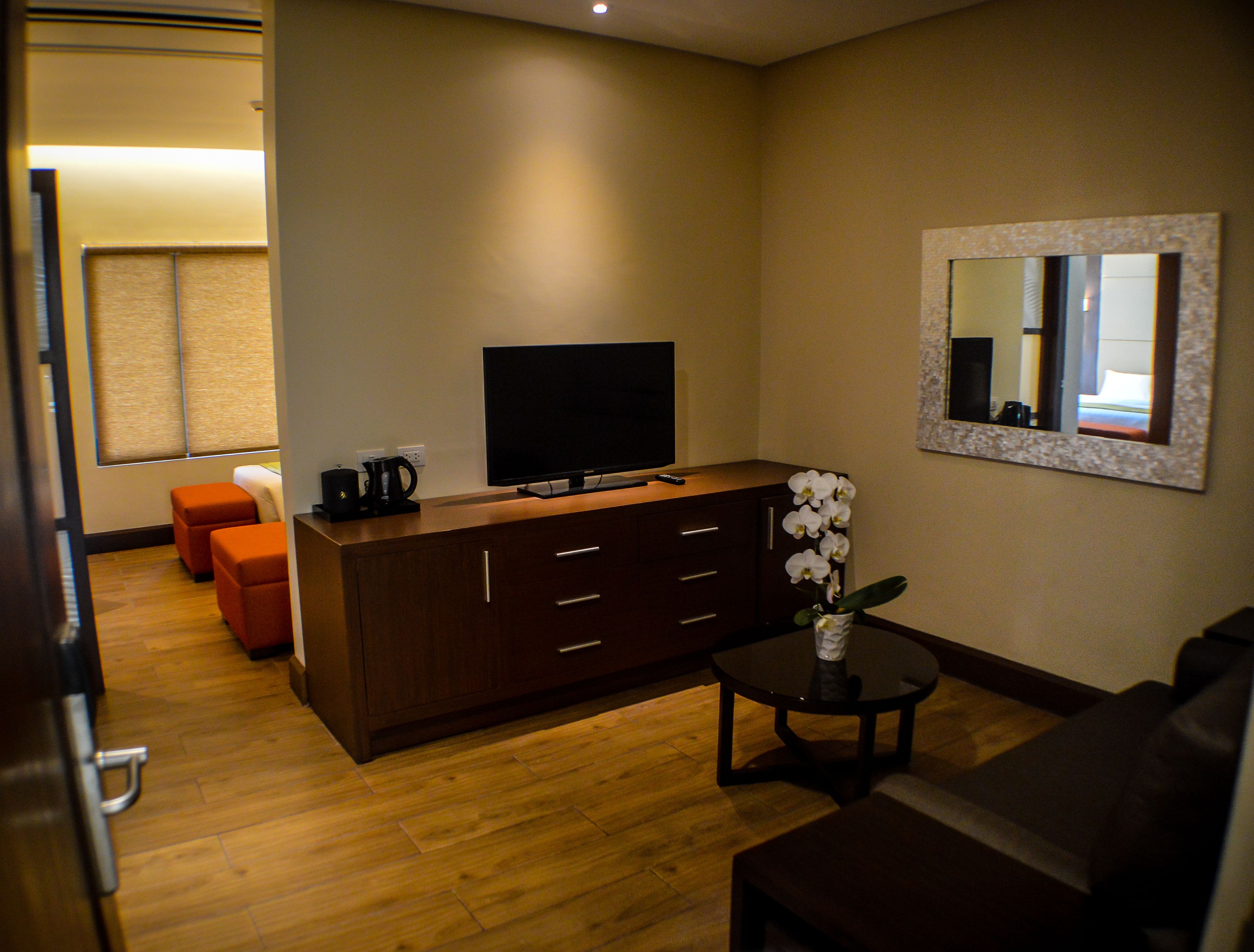 Una habitación de hotel moderna con piso de madera, un sofá marrón oscuro, una pequeña mesa redonda con un jarrón de flores blancas, un televisor de pantalla plana grande sobre un mueble de madera y un espejo en la pared.