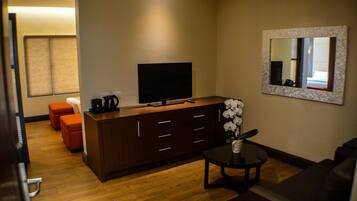 Una habitación de hotel moderna con piso de madera, un sofá marrón oscuro, una pequeña mesa redonda con un jarrón de flores blancas, un televisor de pantalla plana grande sobre un mueble de madera y un espejo en la pared.