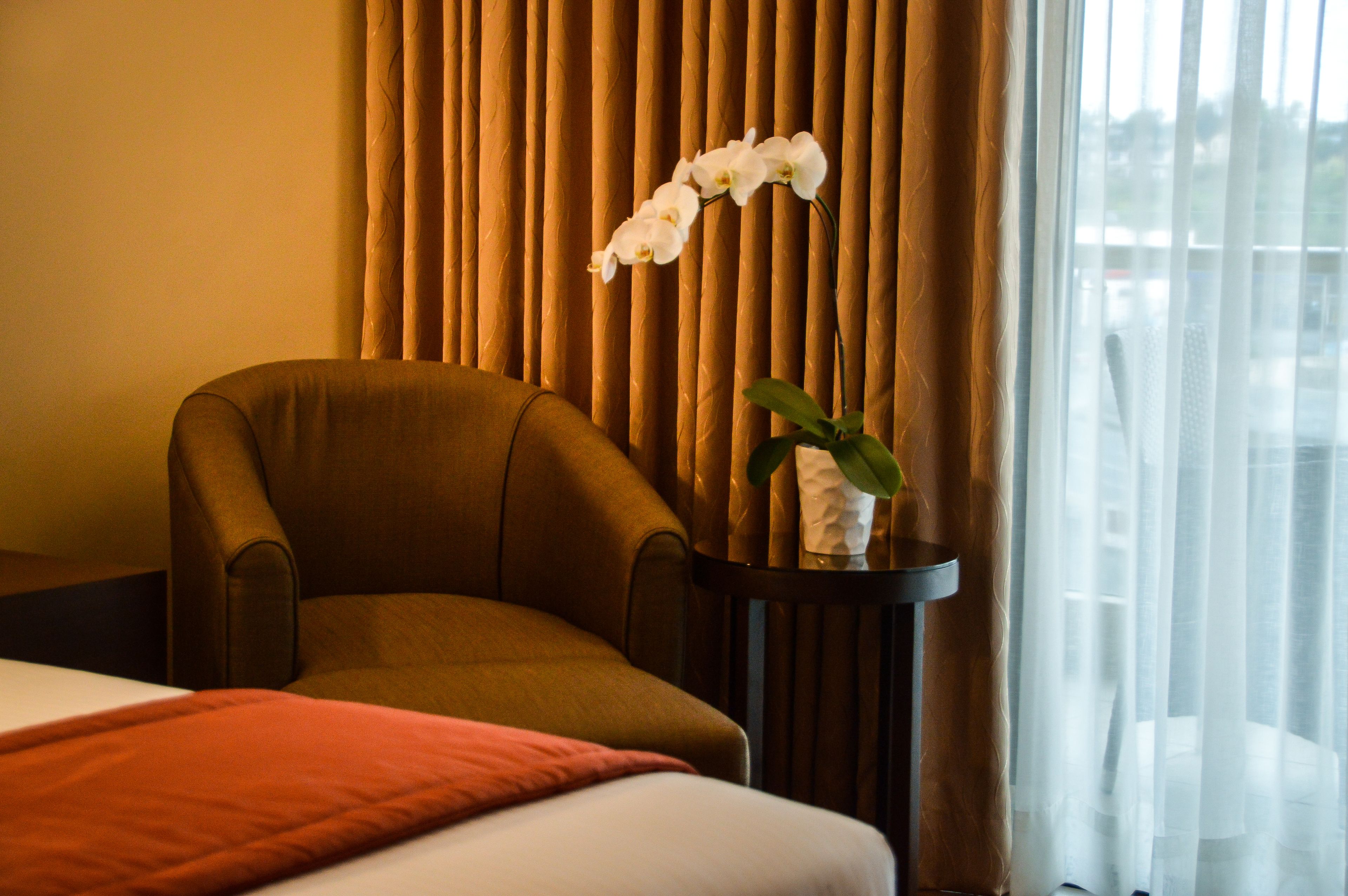 Ein Hotelzimmer mit einem braunen Sessel, einem kleinen Tisch mit einer weißen Orchidee und einem Bett mit roter Decke.
