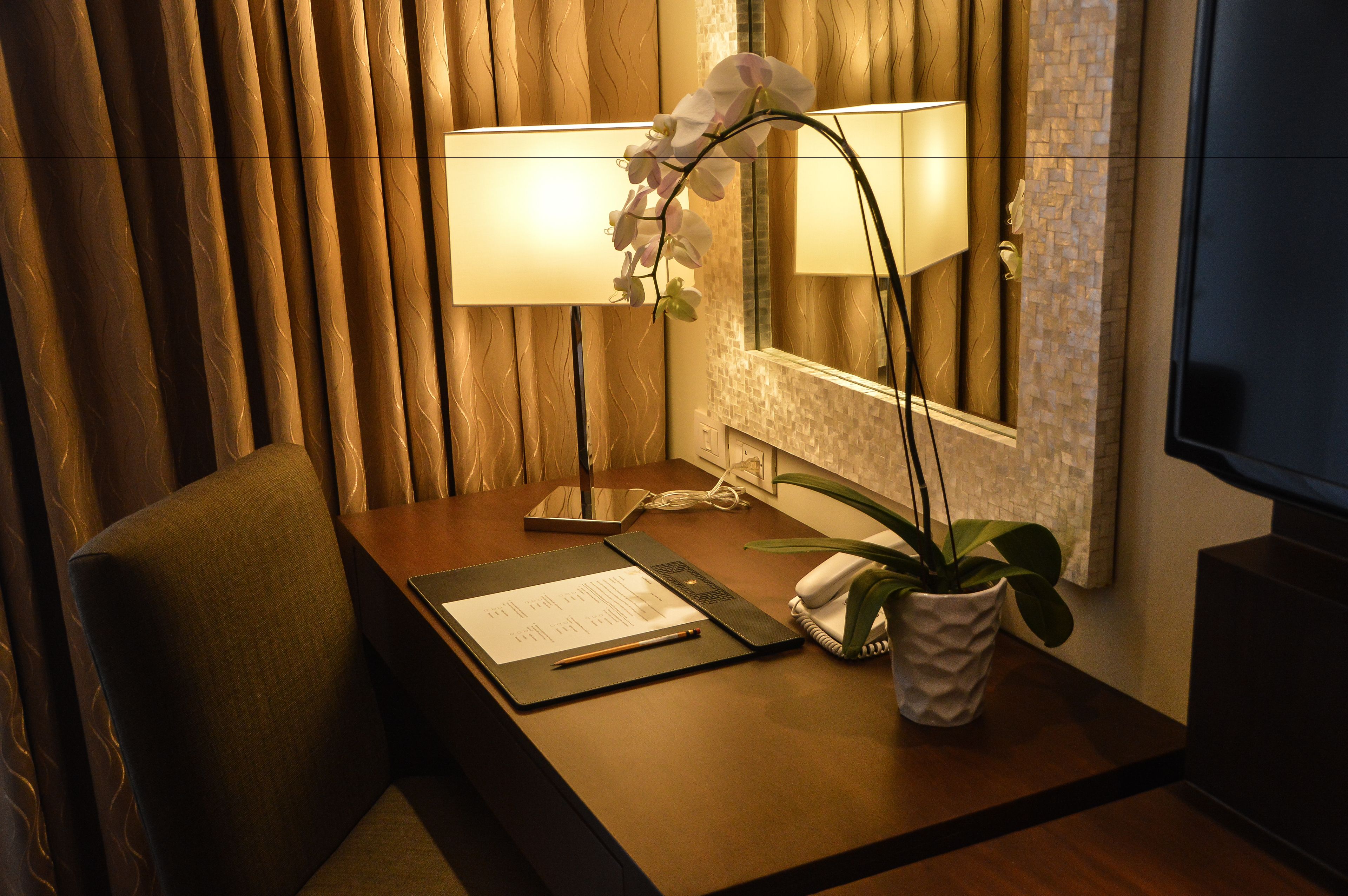 A hotel room desk with a lamp, a telephone, a notepad, a pen, and a potted plant.