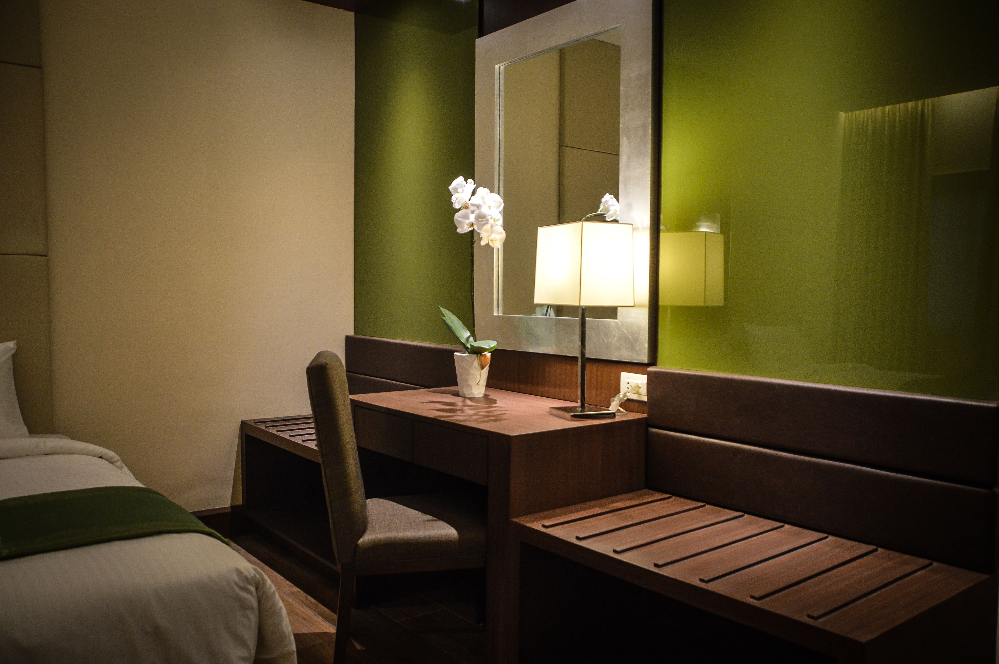 A hotel room with a bed, a desk with a chair, a lamp, and a mirror.