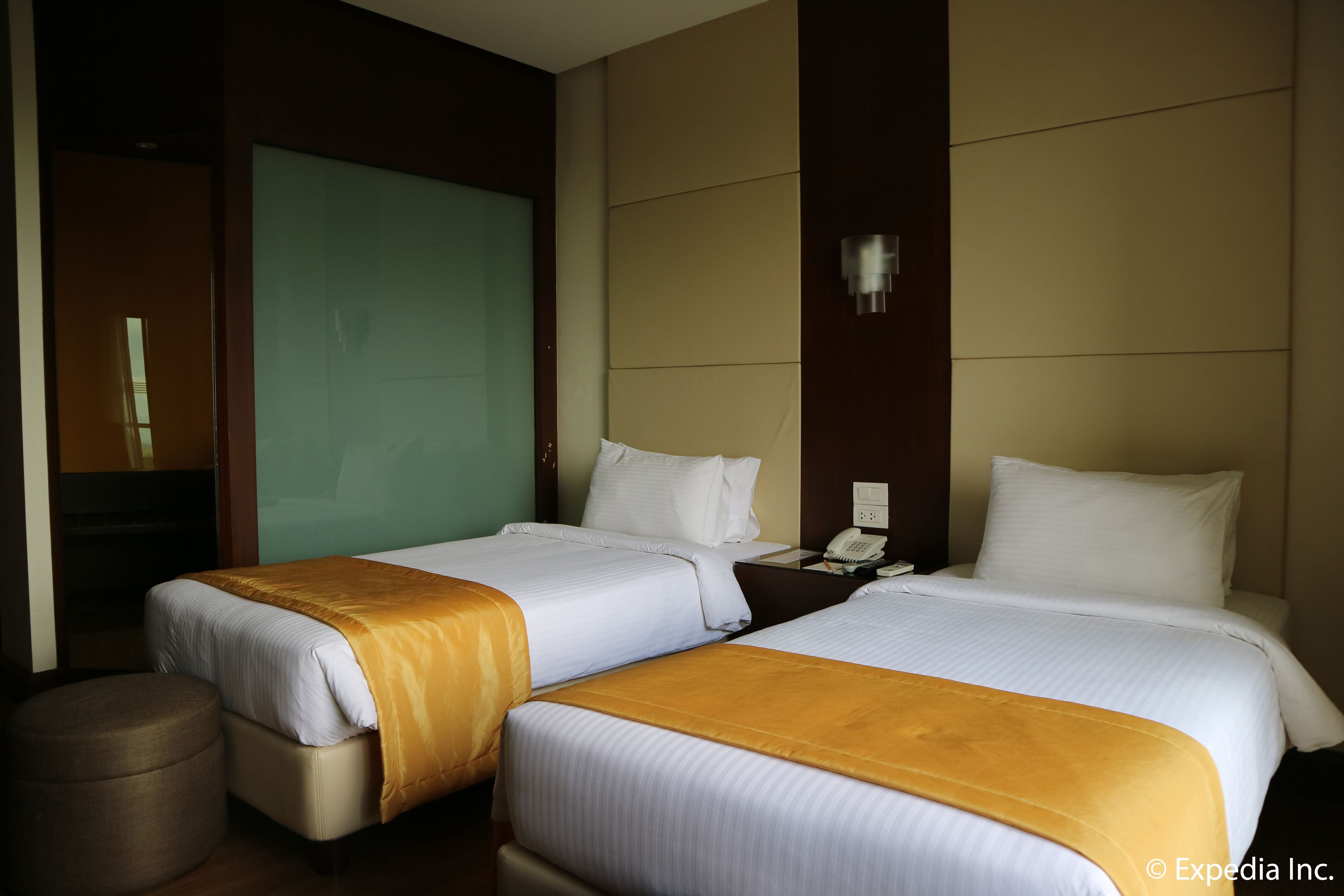 A hotel room with two beds, a small round ottoman, a bedside table with a phone, and a wall-mounted light.