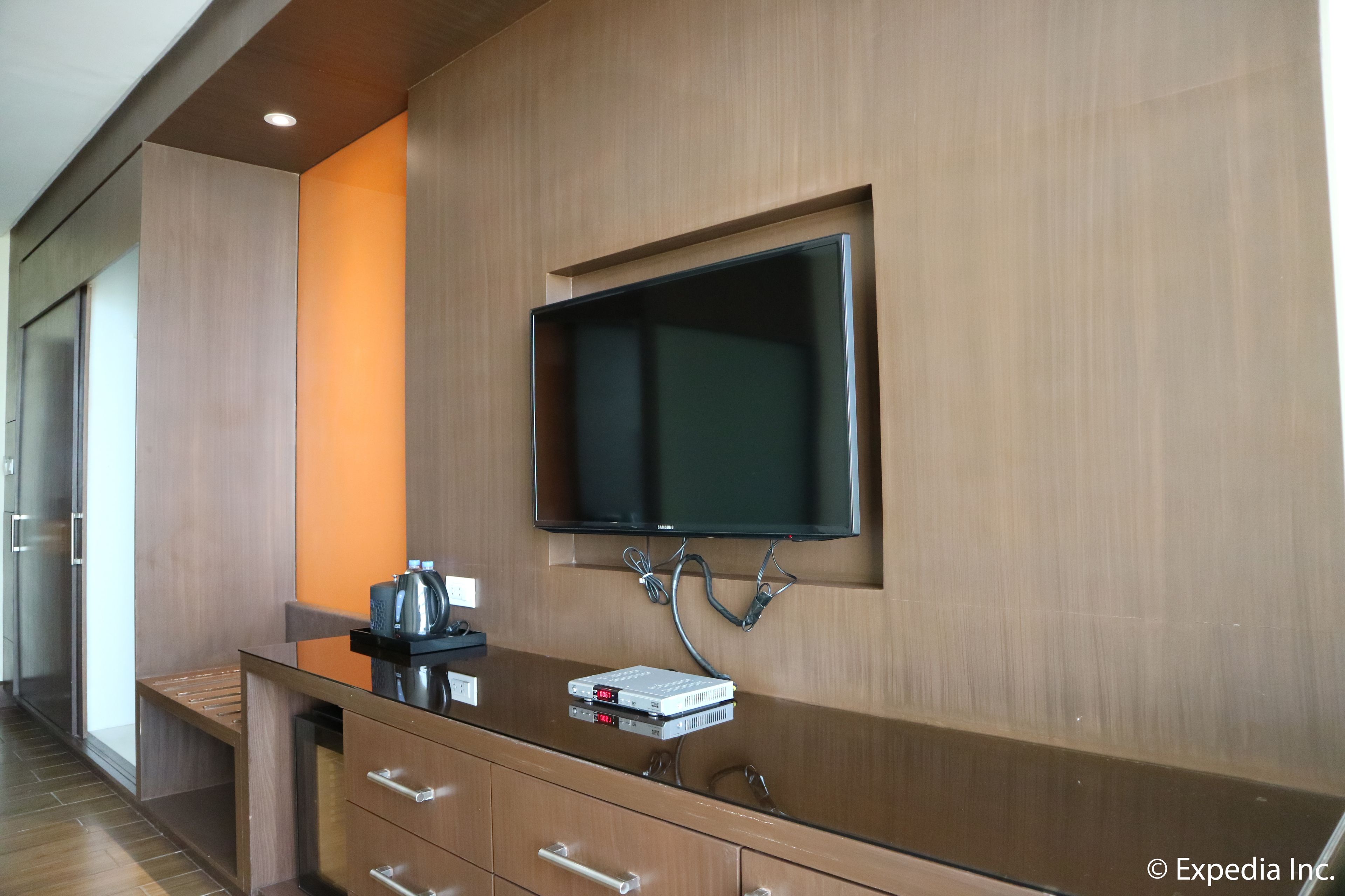A modern hotel room with a wooden vanity, a flat-screen TV mounted on the wall, and a small table with a kettle and a book.