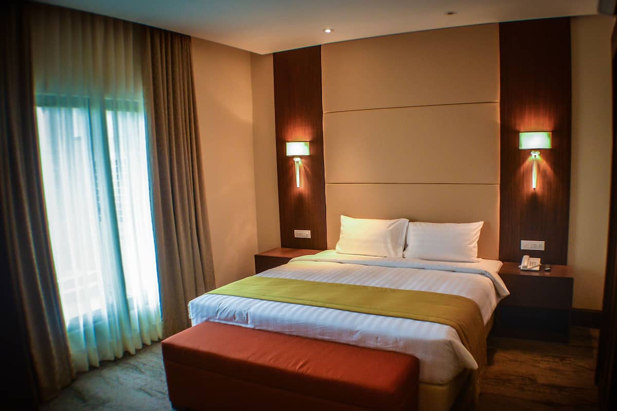 A hotel room with a large bed, a bedside table with a lamp, a telephone, and a window with curtains.
