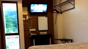 A hotel room with a bed, a television, a desk with a telephone, a mirror, and a window with a view of greenery. - Galaxy Inn (Lumut)