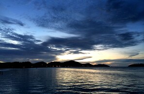 A coastal landscape with a sunset over the water, distant hills, and a city skyline in the background.