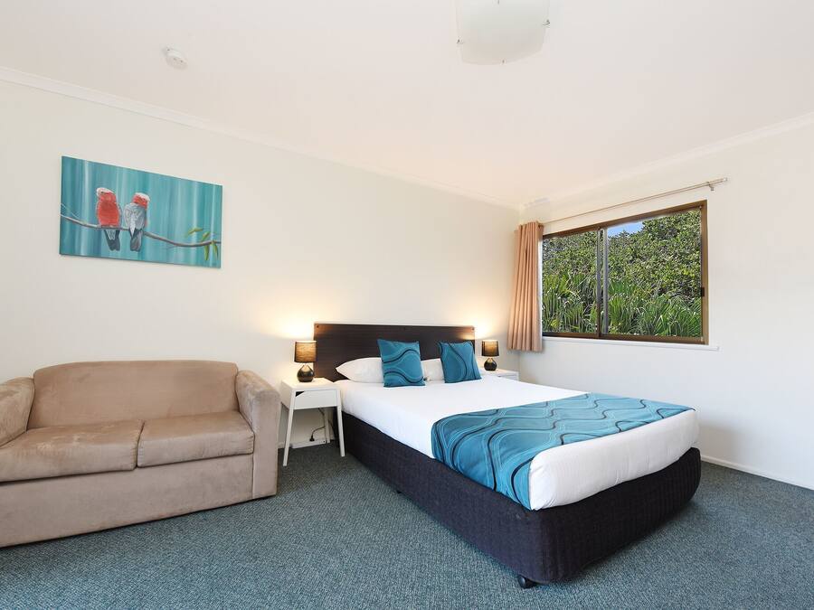 Habitación de hotel con cama, sofá, mesita de noche, lámpara y ventana con cortinas.