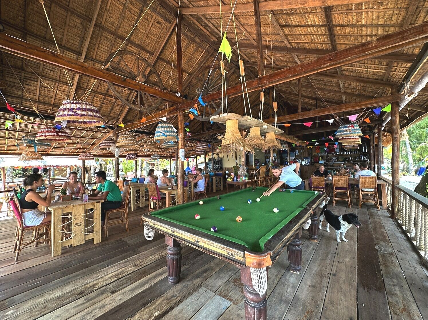 A wooden deck with a pool table, tables, and chairs, under a thatched roof with hanging lamps.
