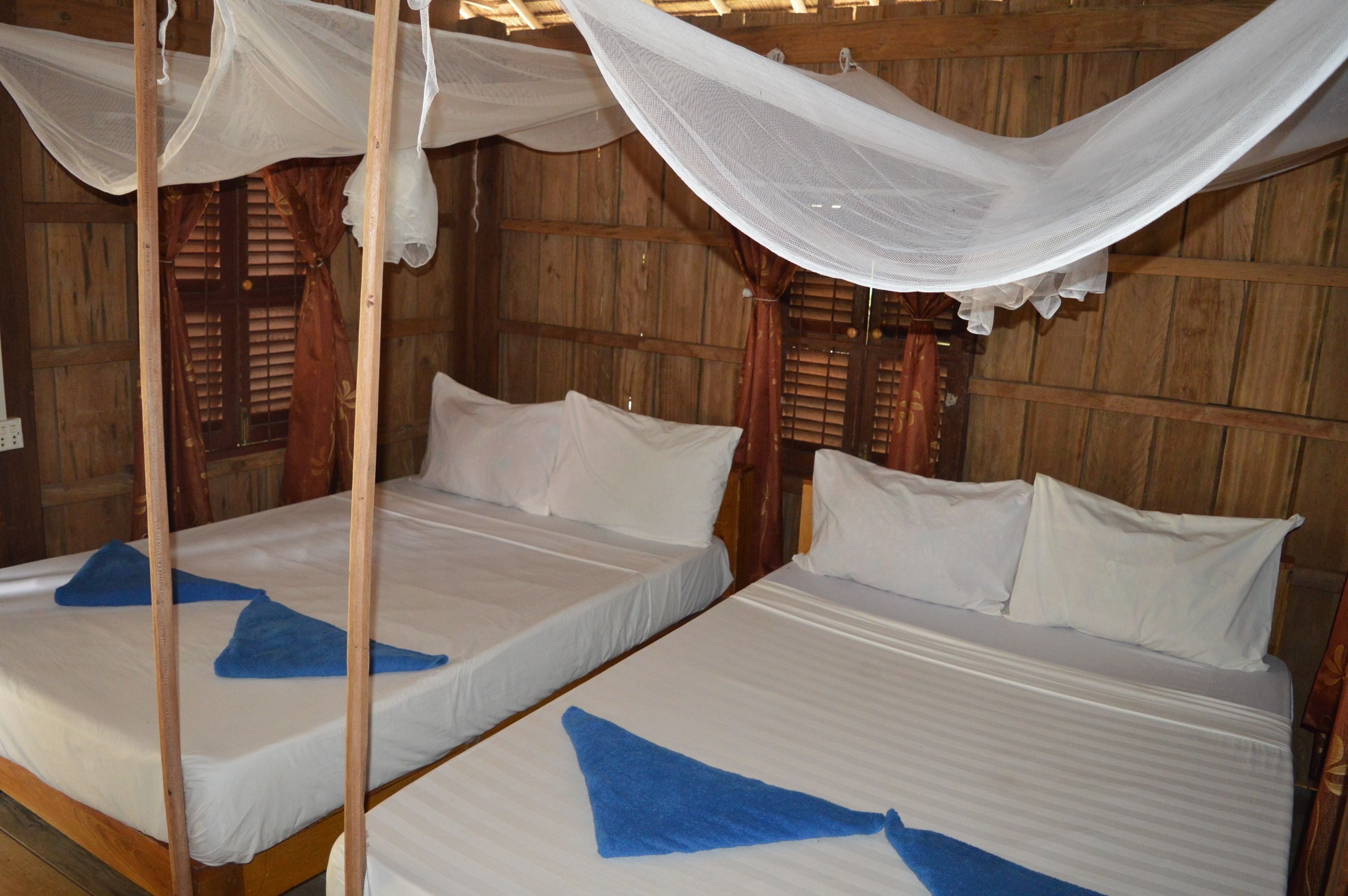 Two single beds with white linens and blue towels, wooden walls, and a canopy bed with white netting.