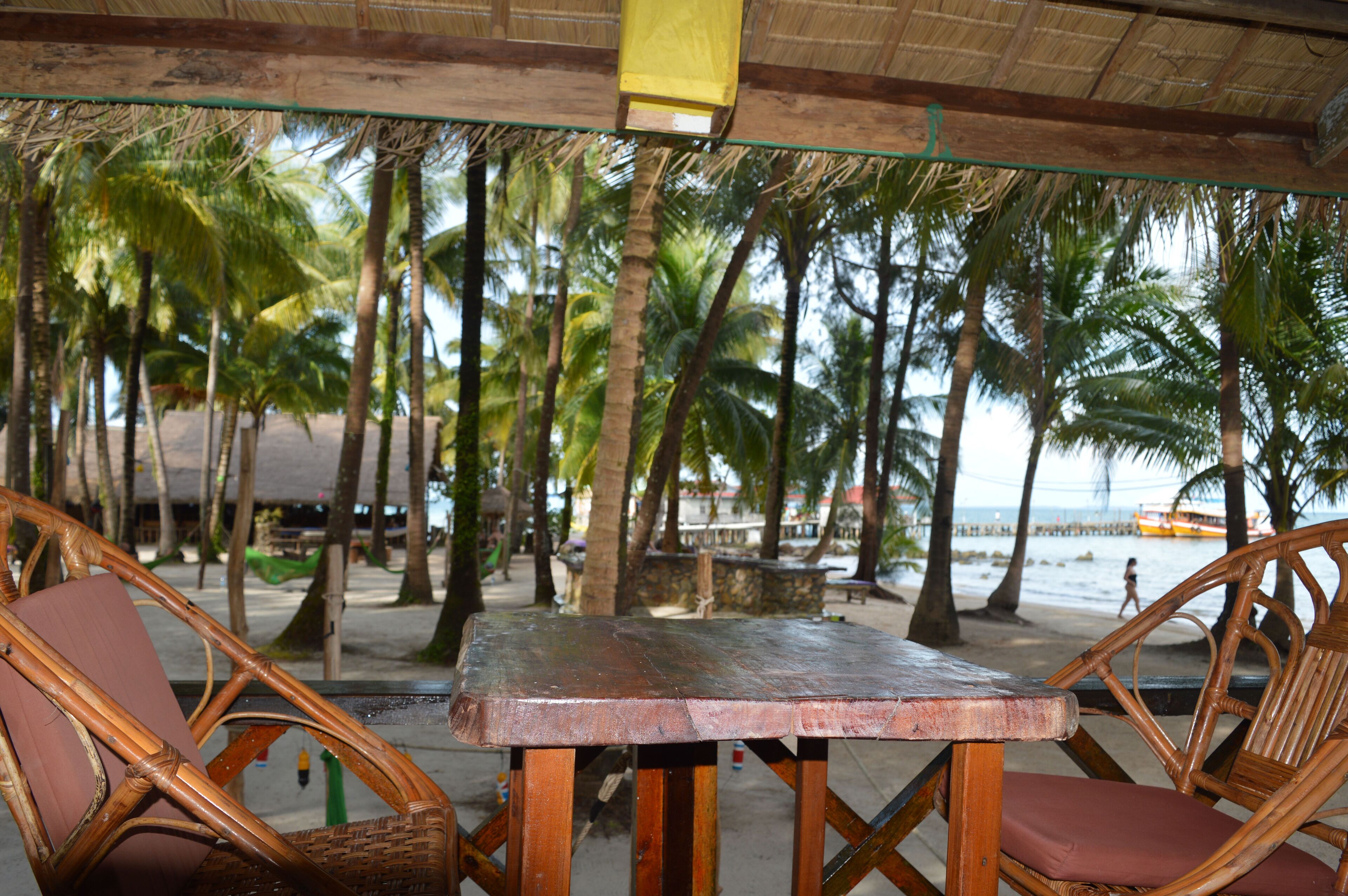 A beachside seating area with a table and chairs, surrounded by palm trees and a view of the beach.