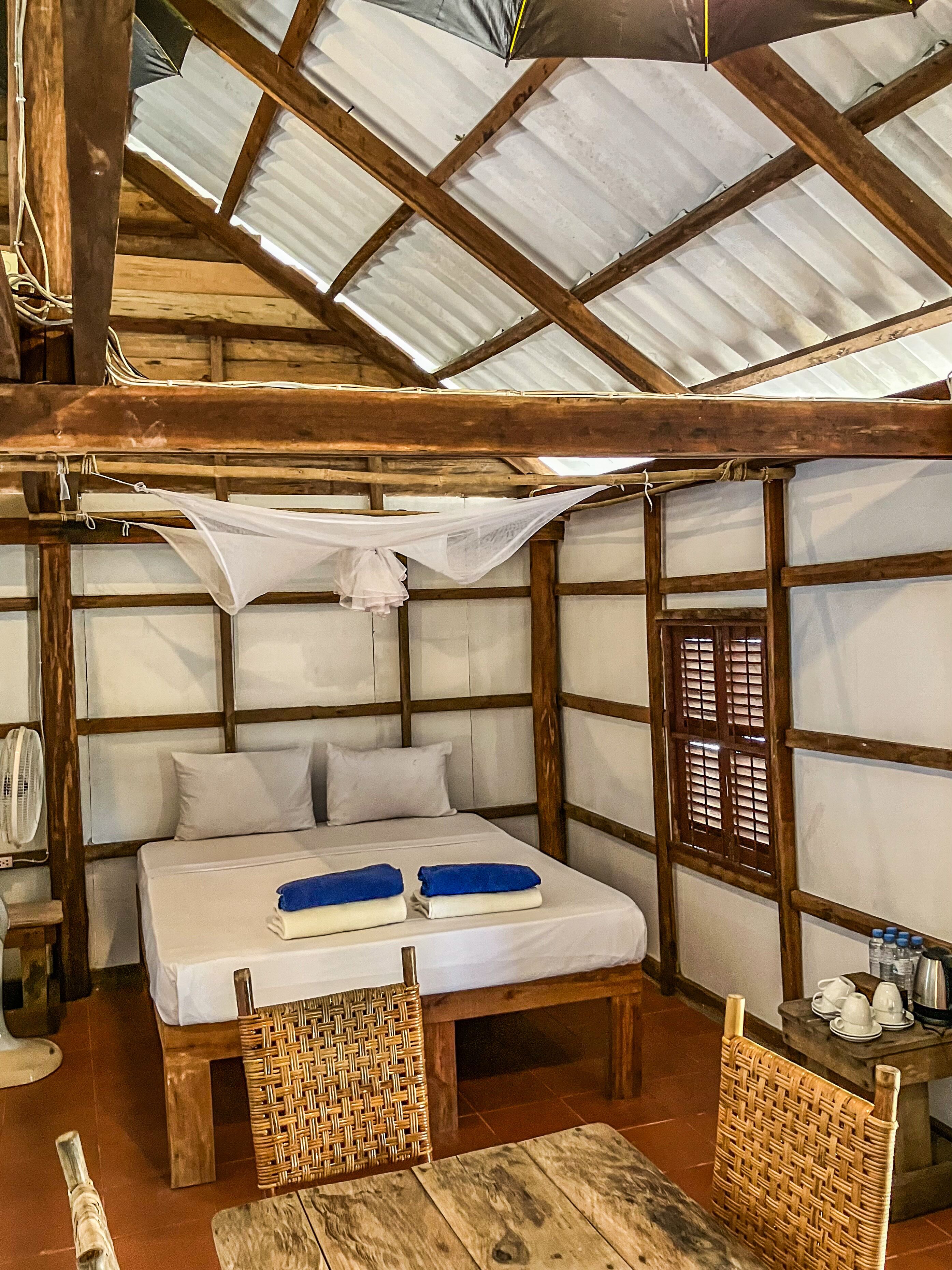 Een eenvoudige kamer met een bed, twee geweven stoelen, een tafeltje en een ventilator.