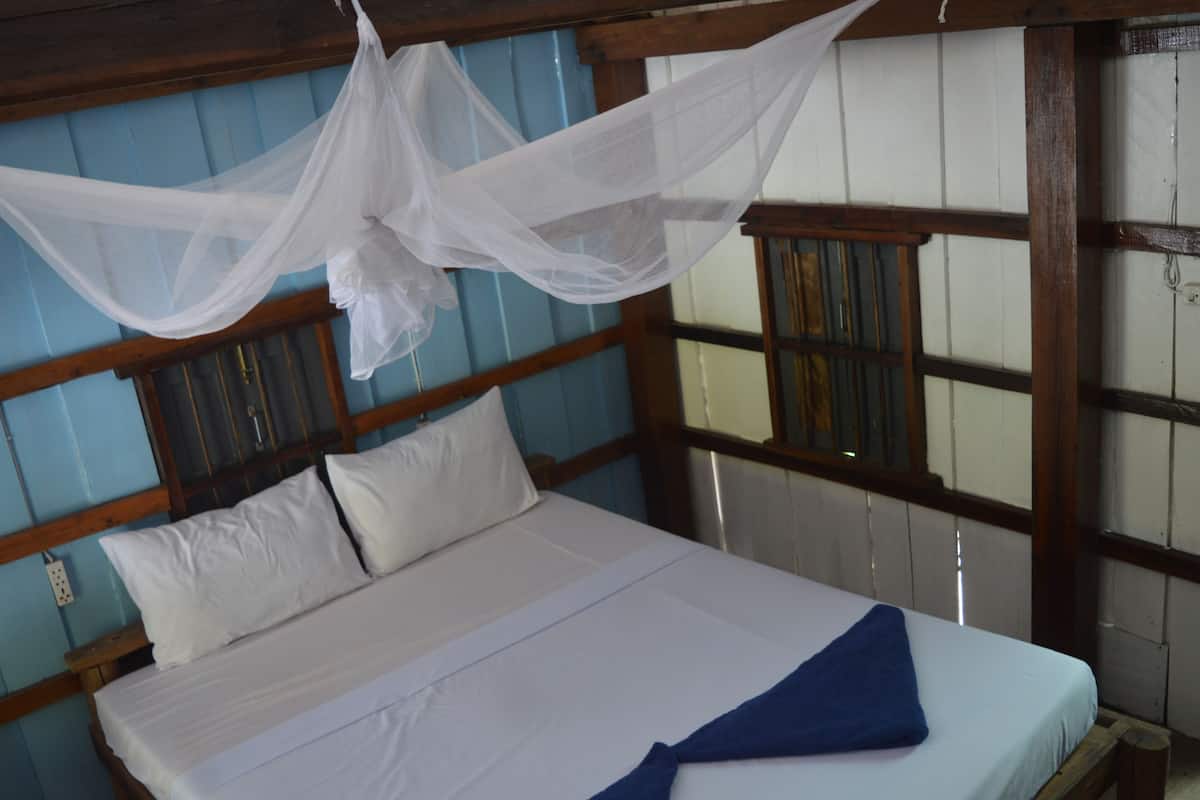 Un lit avec du linge de lit blanc et une serviette bleue, une tête de lit en bois et une moustiquaire au-dessus.
