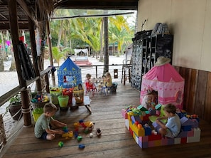 Une terrasse en bois où des enfants jouent avec des jouets, une petite maison de jeu bleue et une tente rose.