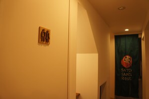 A hallway with a wooden sign indicating a men's restroom, a green banner with a logo and text, and a white door at the end. - Sato san's Rest - Hostel (Tokyo)