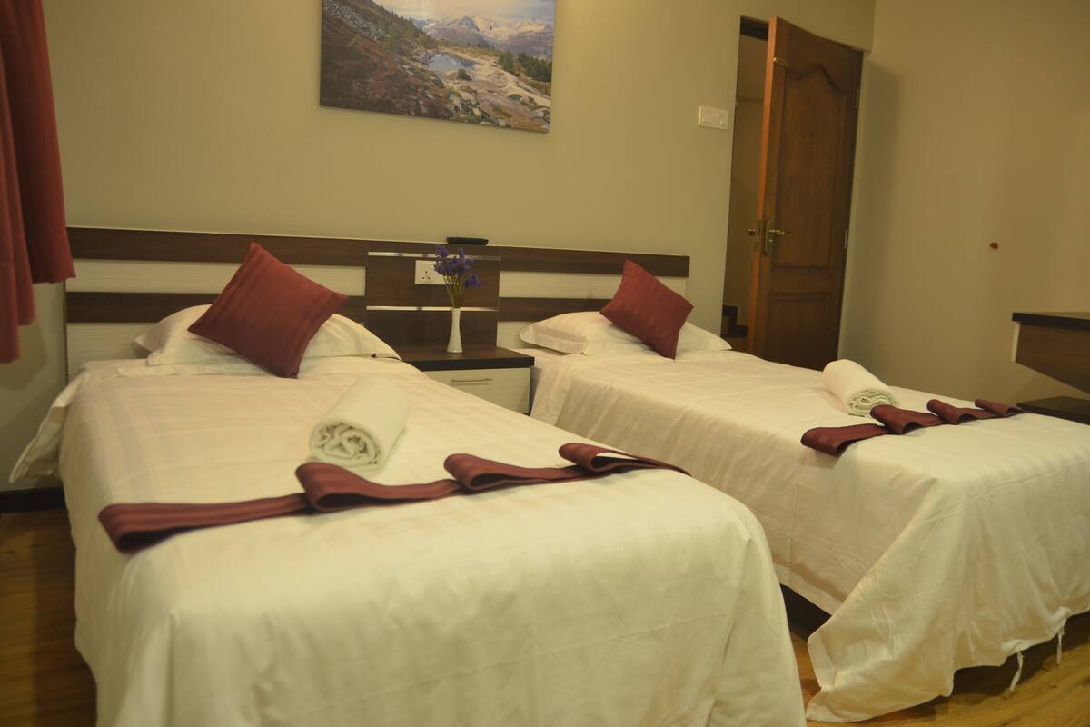 Two single beds with white linens and red pillows, a wooden headboard, a nightstand with a vase of flowers, and a painting of a mountainous landscape on the wall.