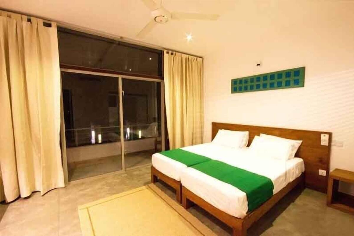 Una habitación de hotel con una cama grande, un ventilador de techo, una ventana con cortinas y un adorno verde en la pared.