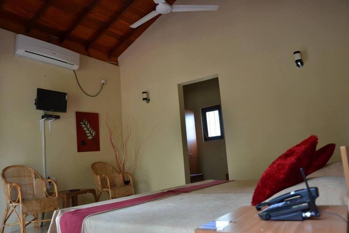 Un dormitorio con cama, ventilador de techo, almohada roja, mesita de noche, lámpara y televisor montado en la pared.