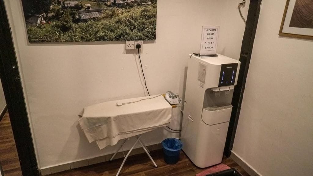 A room with a small ironing station, a water dispenser, and a trash bin.