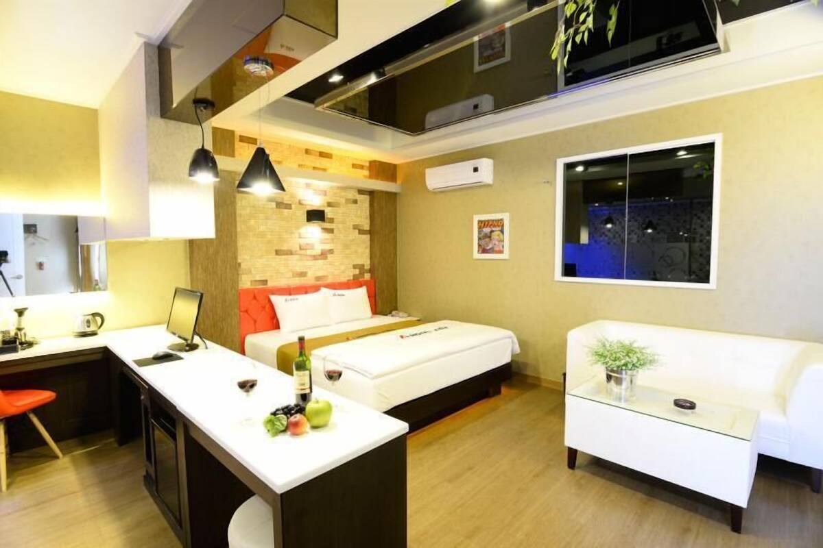 A modern hotel room with a kitchenette, a bed with a red headboard, a white sofa, and a large window.