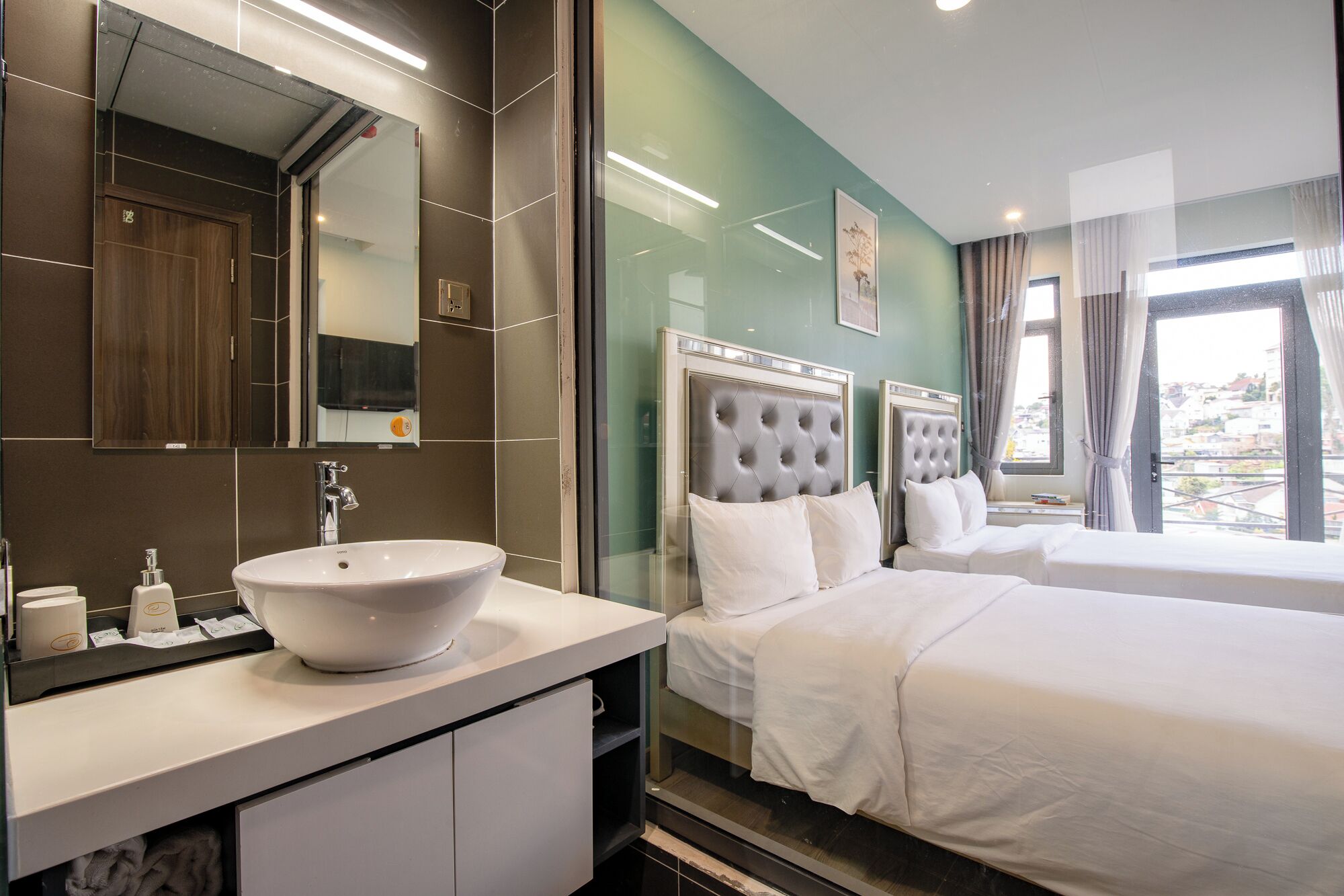 A hotel room with a large white sink, a mirror, a bed with a tufted headboard, and a window with curtains.