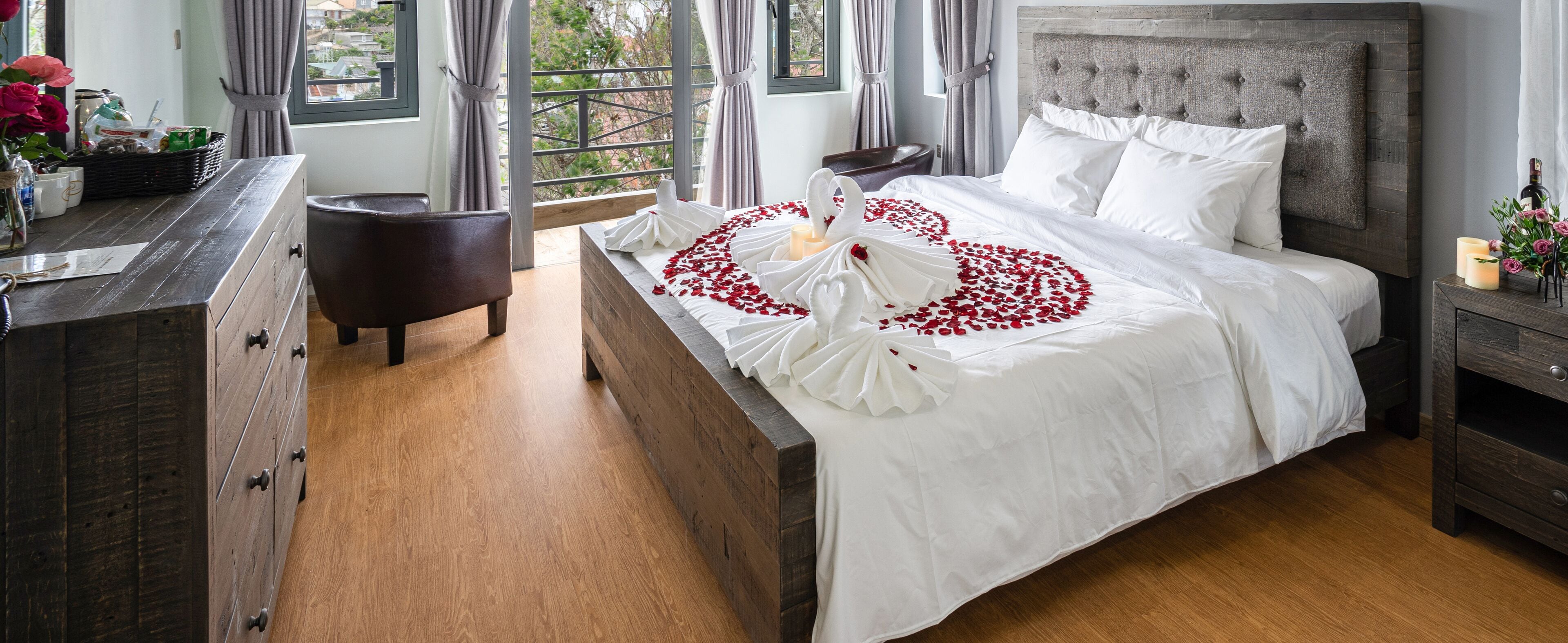 A neatly made bed with white linens and red rose petals arranged in a heart shape, a wooden dresser with drawers, a small round table, and a nightstand with candles.