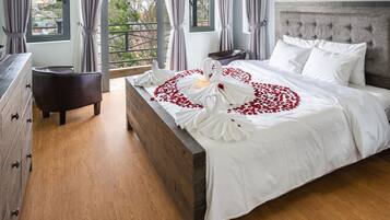 A neatly made bed with white linens and red rose petals arranged in a heart shape, a wooden dresser with drawers, a small round table, and a nightstand with candles.
