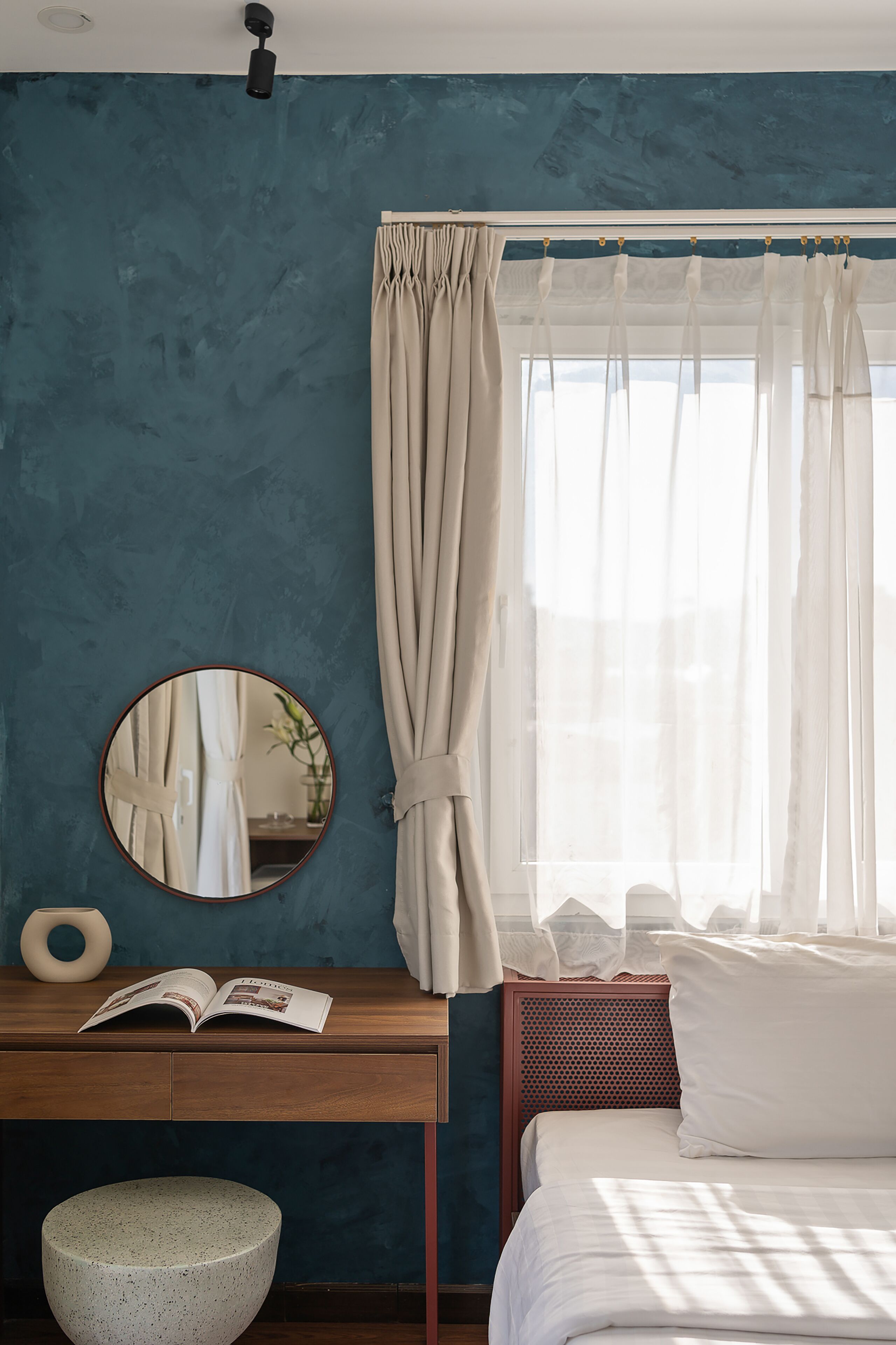Una camera da letto con un letto, un comodino con un libro, uno specchio rotondo e una finestra con tende.