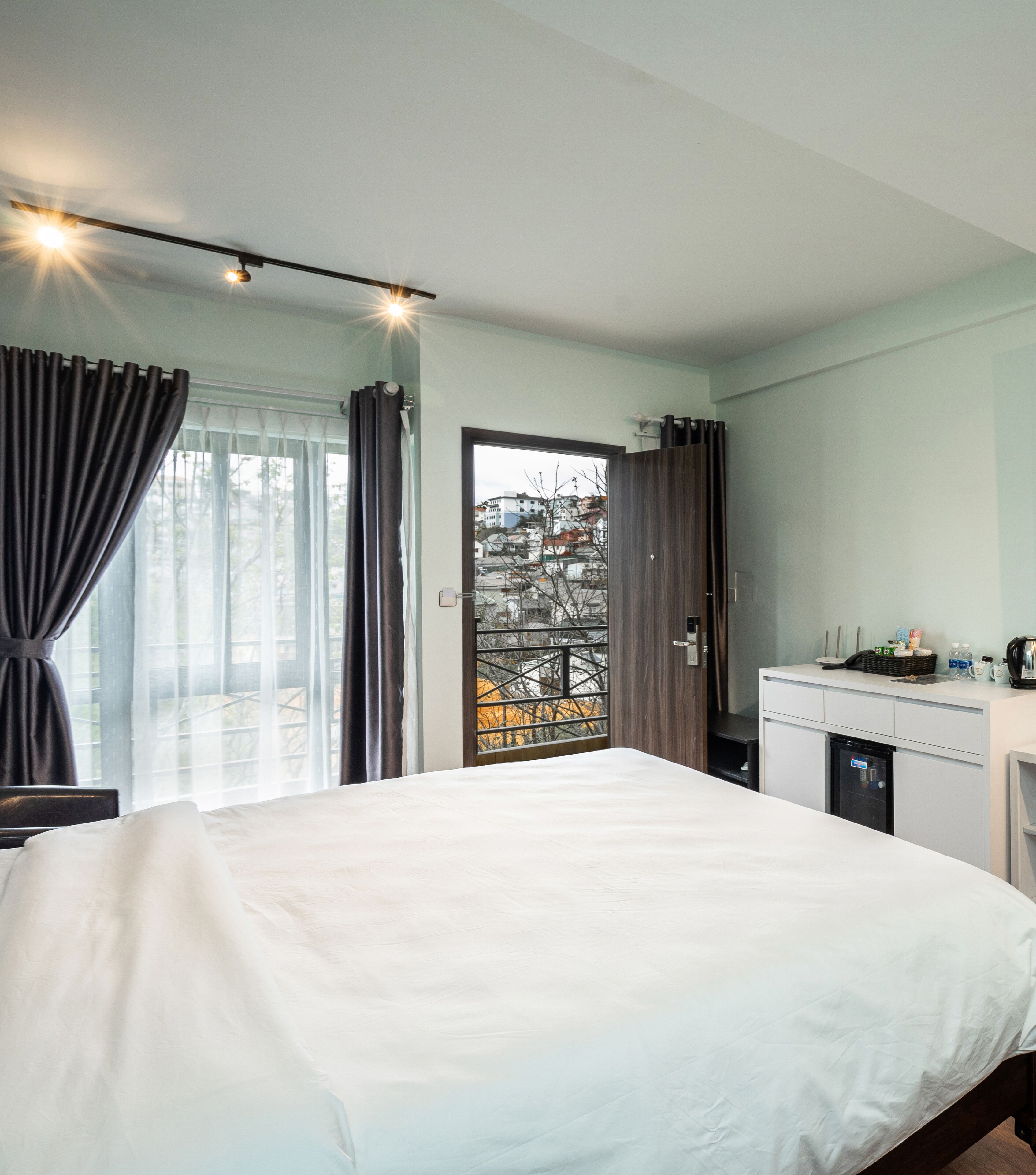 A hotel room with a bed, a desk with a computer, a small refrigerator, and a window with curtains.