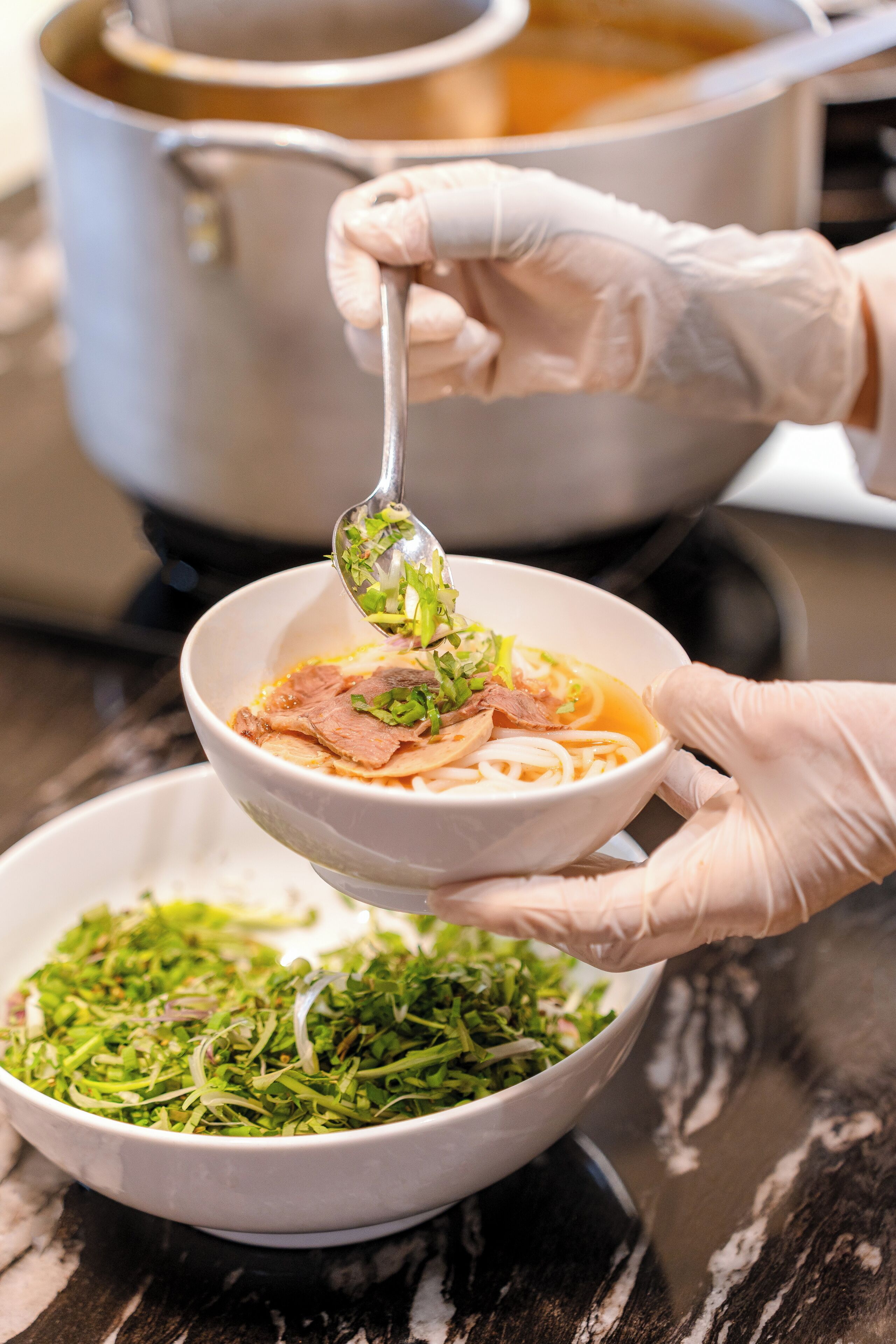 A person holding a bowl of soup with noodles and meat, with a pot of soup in the background.