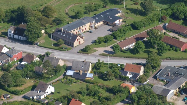 Eine Vogelperspektive zeigt eine Wohngegend mit einem großen Gebäude, vermutlich ein Gemeinschaftszentrum oder ein Vereinshaus, umgeben von kleineren Häusern und einer Straße.