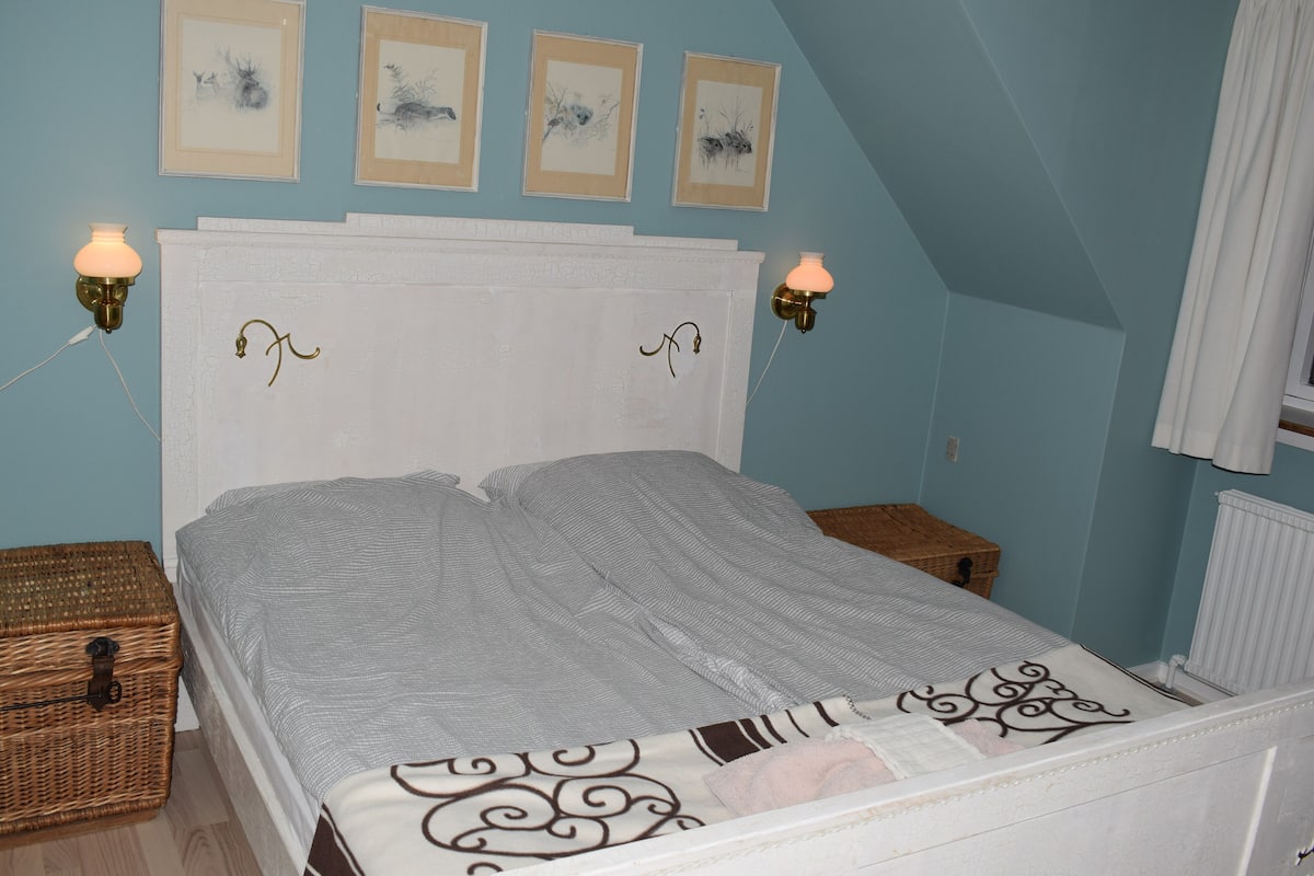 A bedroom with a white headboard, two bedside tables, a bed with a striped bedspread, and wall-mounted sconces.
