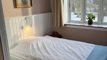A single bed with a white mattress, a bedside table, a small potted plant on the windowsill, and a framed picture on the wall.
