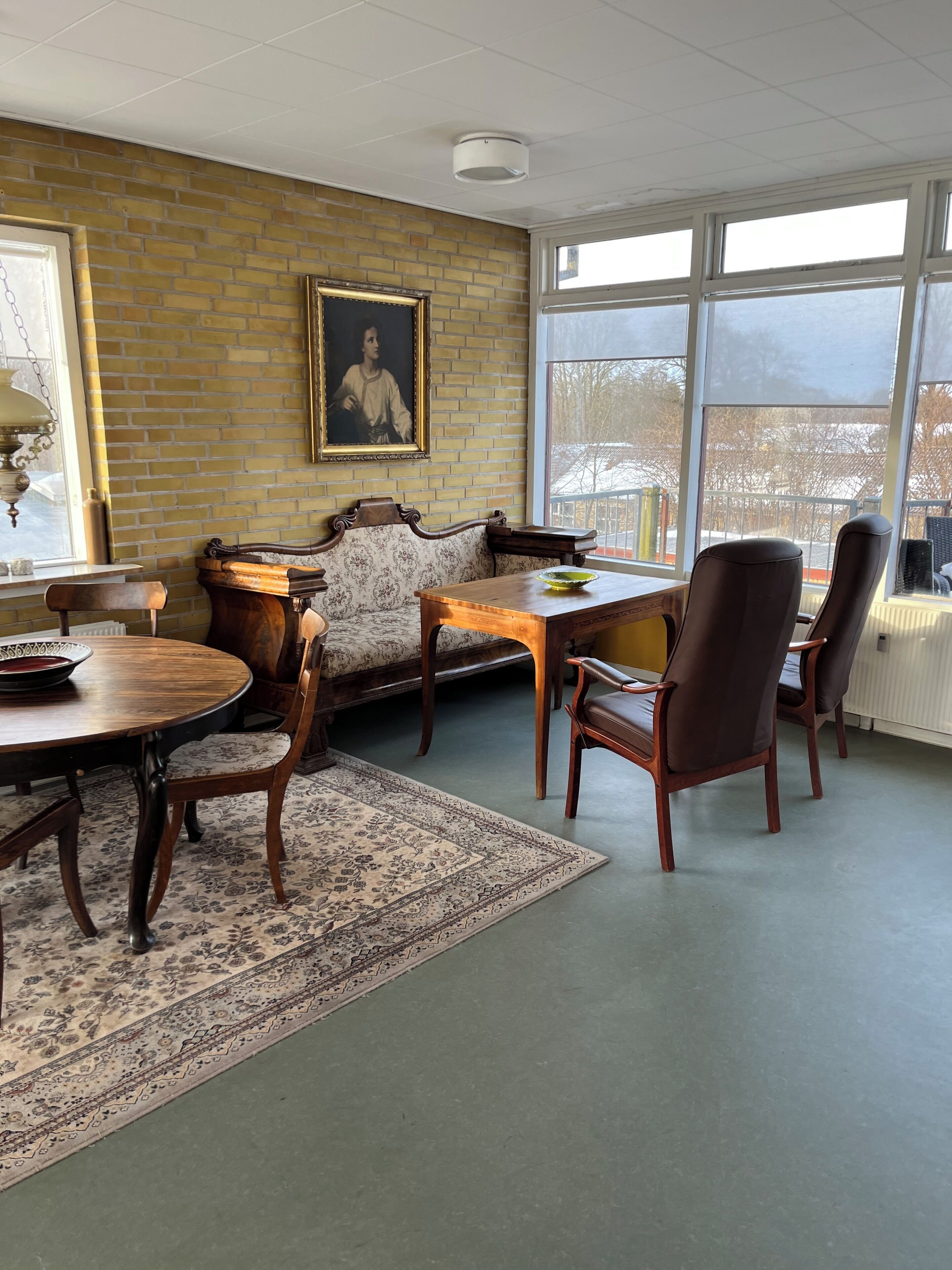 A room with a brick wall, a framed portrait, a wooden table with chairs, and a large window.
