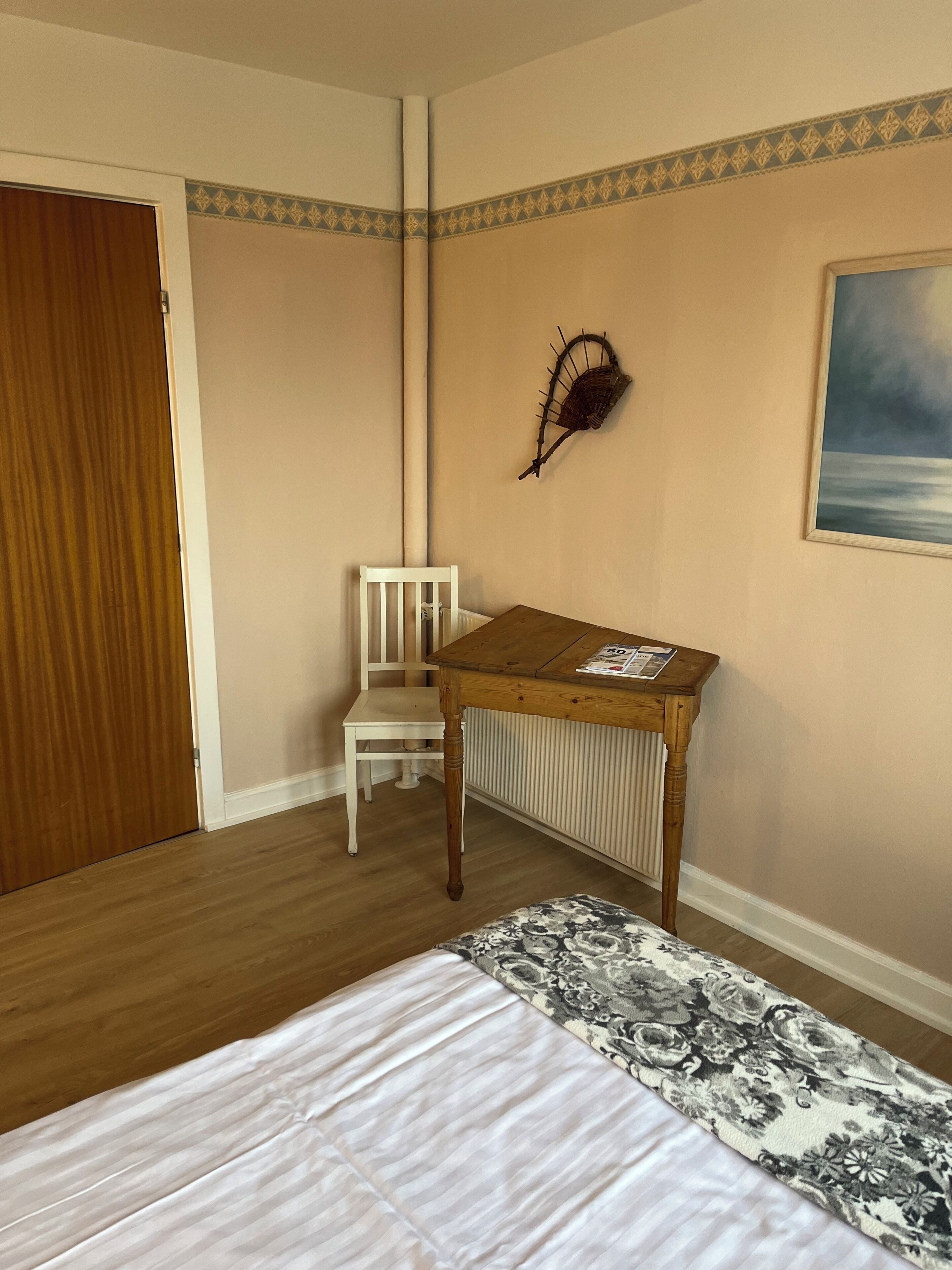 A bedroom with a bed, a wooden table, a chair, a radiator, a door, and a wall decoration.