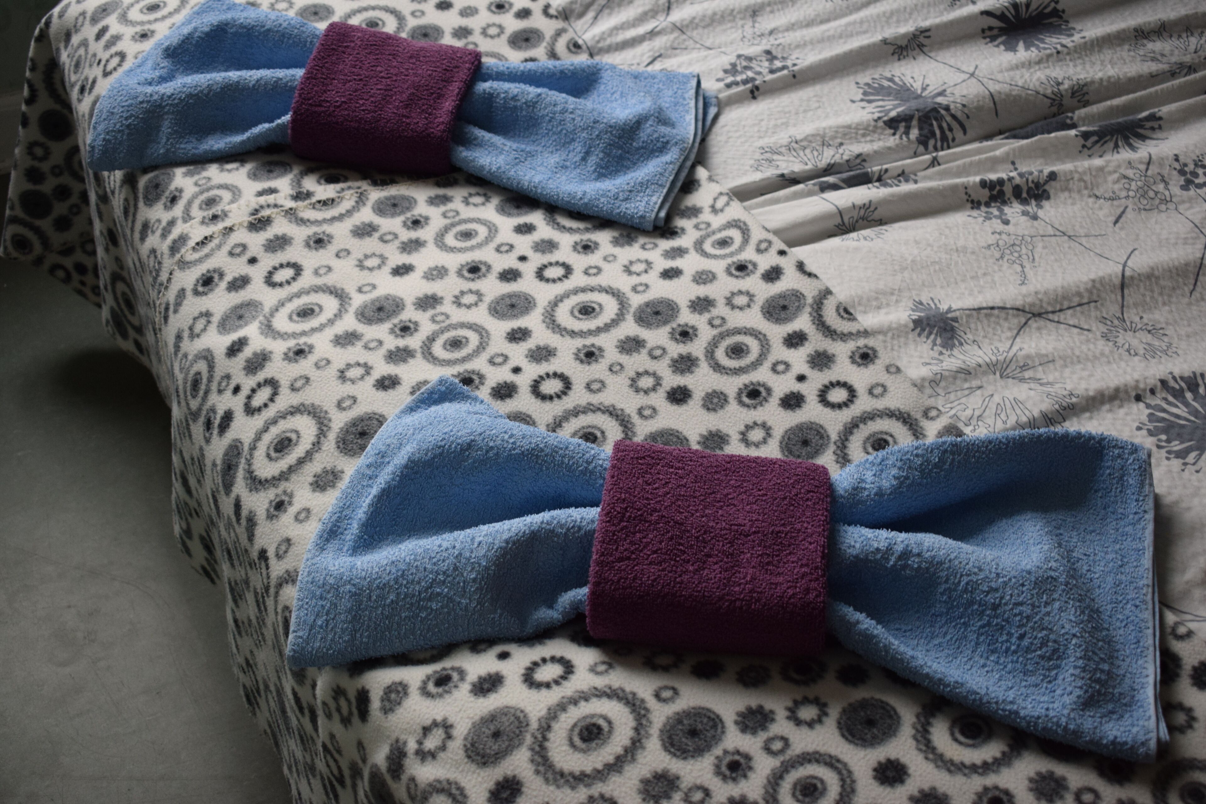 A bed with a patterned bedspread and two towels tied with blue and purple ribbons.