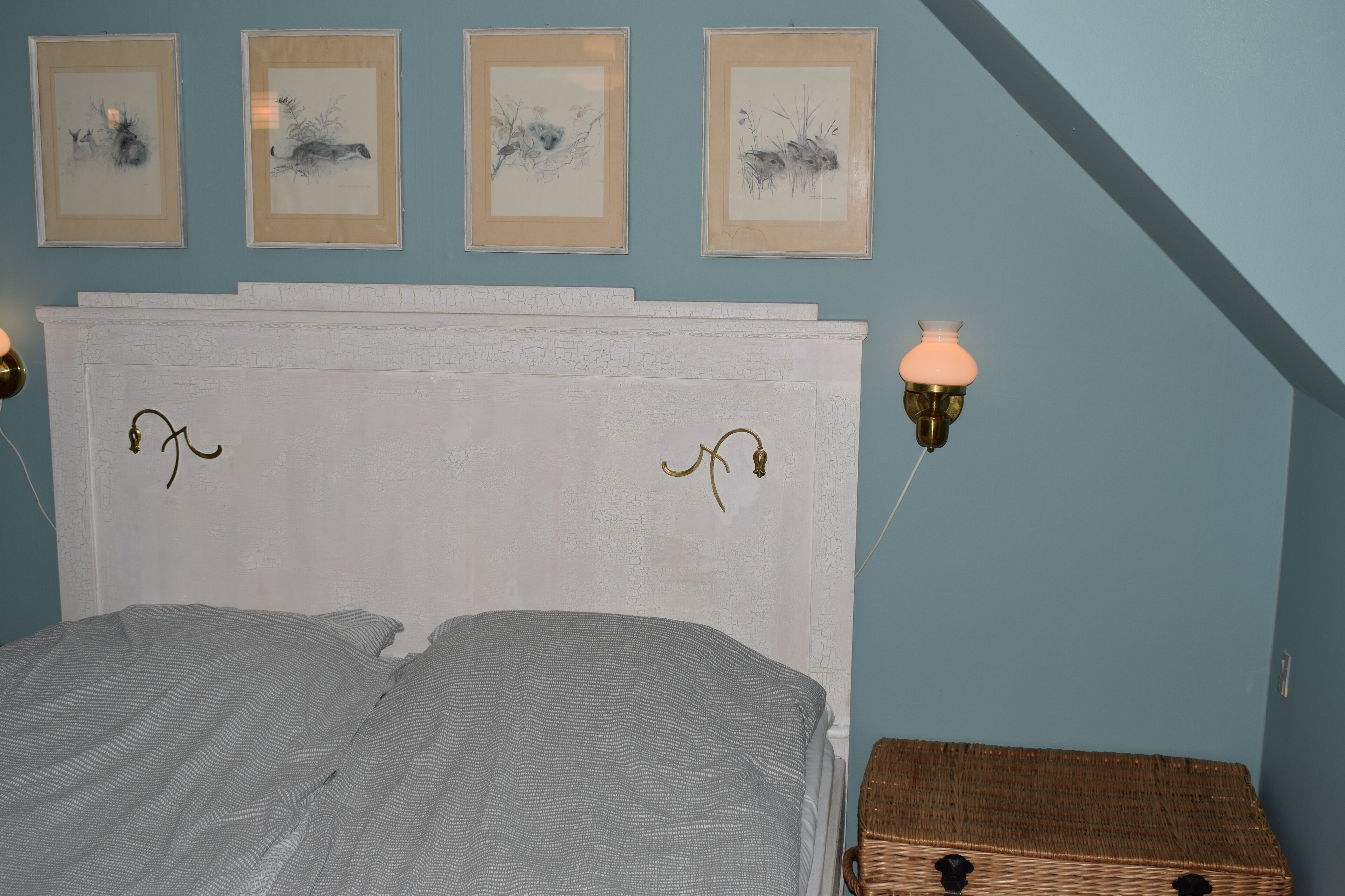 A bedroom with a white headboard, four framed artworks on the wall, a bedside lamp, and a wicker chest of drawers.