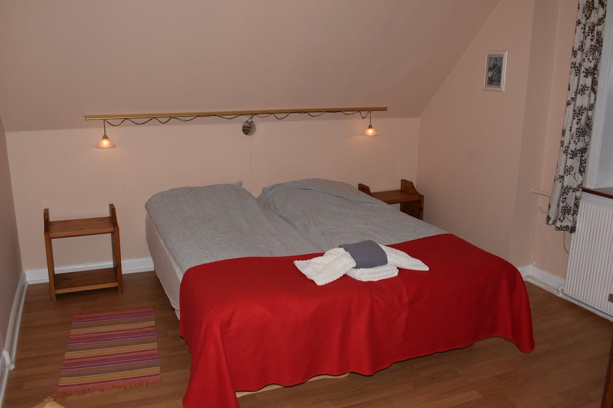 A bedroom with a bed, a small wooden nightstand, a red bedspread, a striped rug, and a ceiling light fixture.