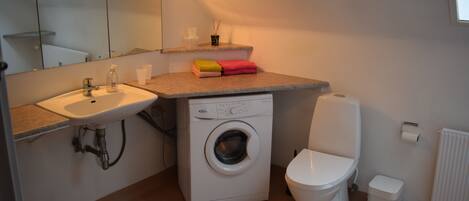 A compact bathroom with a sink, washing machine, toilet, and a mirror.